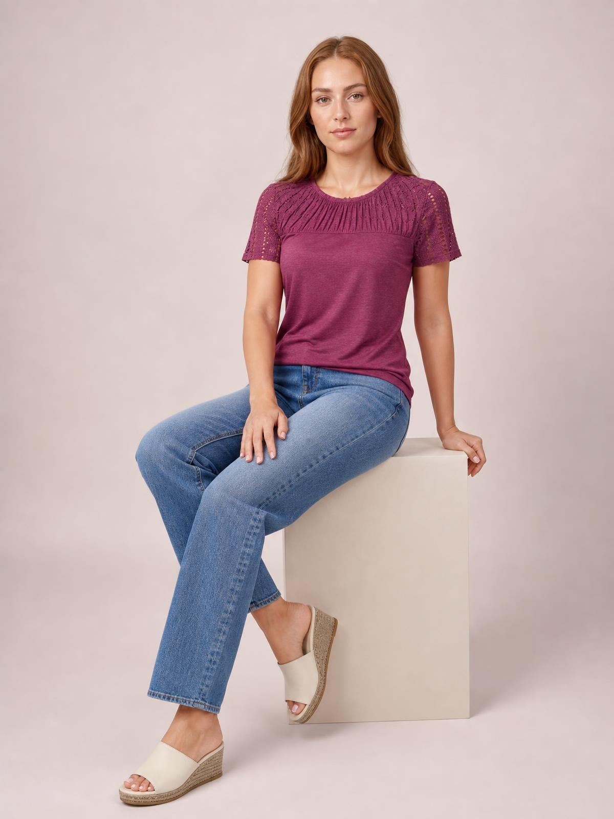 A woman sits in a relaxed pose wearing a burgundy smocked round-neck short-sleeve T-shirt with cutout detailing and semi-sheer sleeves.