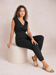 A model sits in a black smocked sleeveless V-neck jumpsuit with a fitted waist, relaxed tapered legs, and visible side pockets.