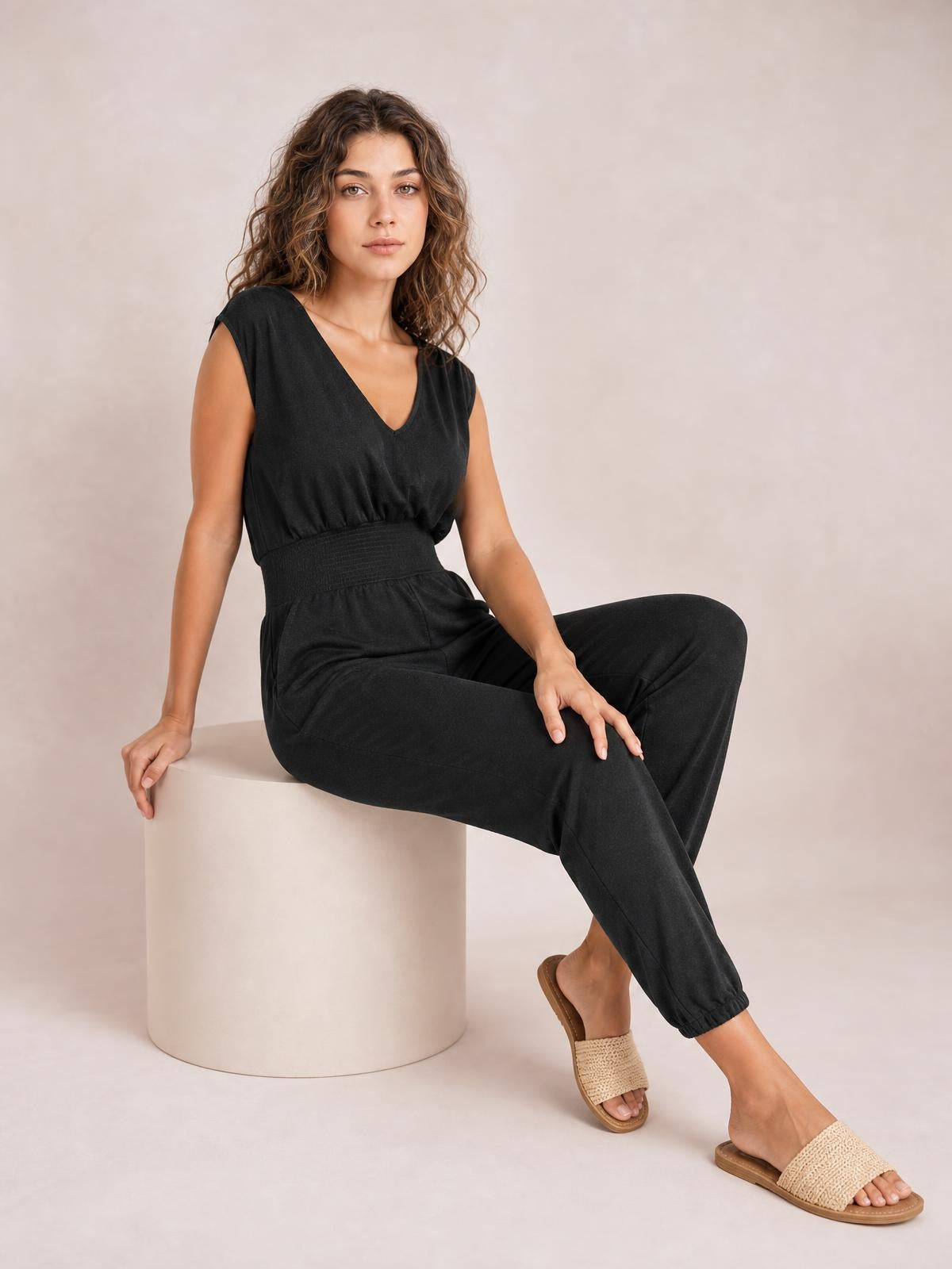 A model sits in a black smocked sleeveless V-neck jumpsuit with a fitted waist, relaxed tapered legs, and visible side pockets.