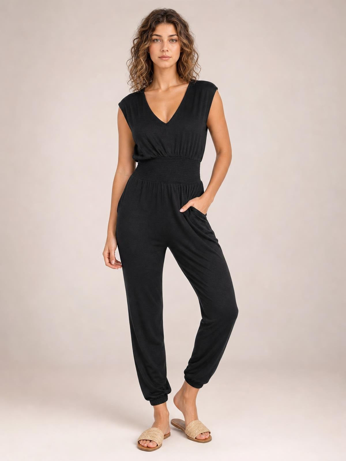 Model wearing a black smocked sleeveless V-neck jumpsuit with a fitted waist, tapered legs, and convenient side pockets, posing with one hand tucked inside.