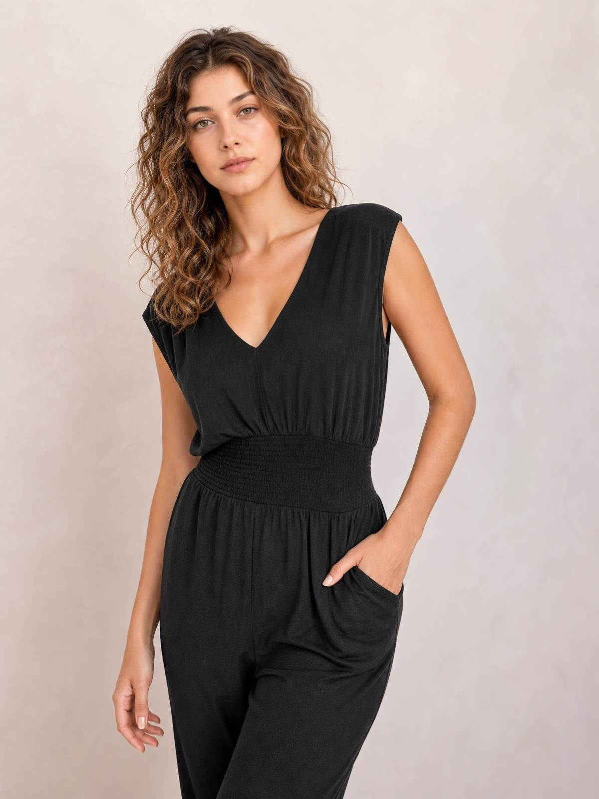 A model poses in a black sleeveless V-neck jumpsuit with a smocked waist, relaxed wide-leg silhouette, and one hand tucked into a pocket.