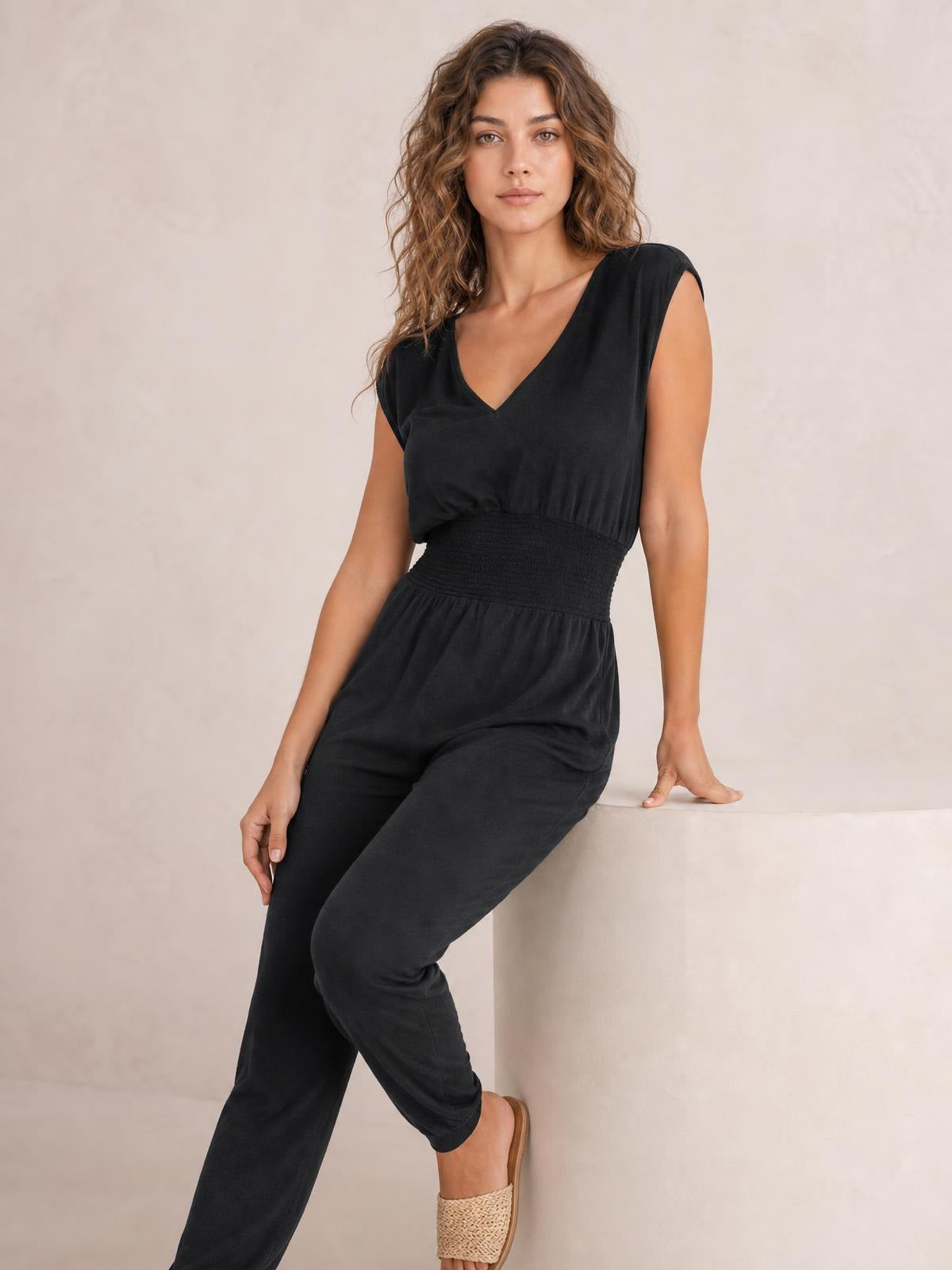 Model wearing a black smocked sleeveless V-neck jumpsuit with a relaxed straight-leg silhouette, seated with one leg crossed.