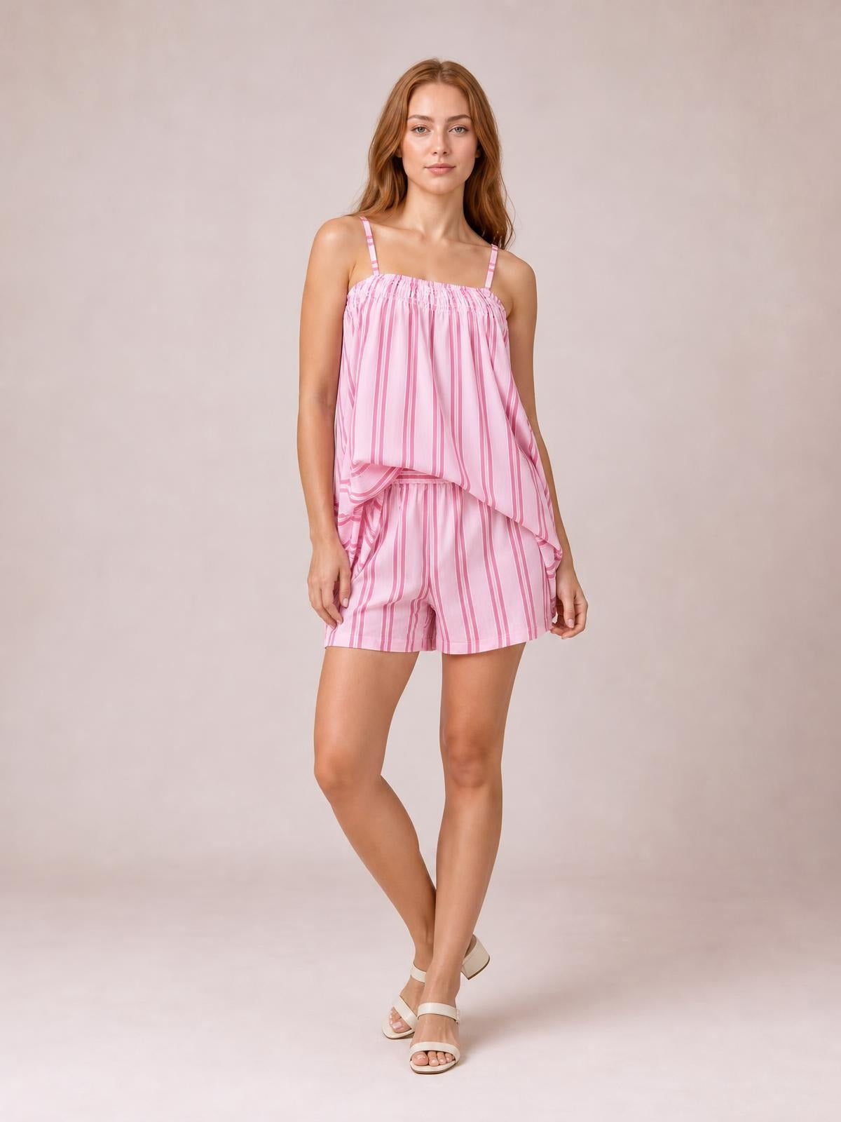 A model poses with crossed legs in a pink striped smocked square-neck cami and matching loose-fit shorts with thin straps and a relaxed silhouette.