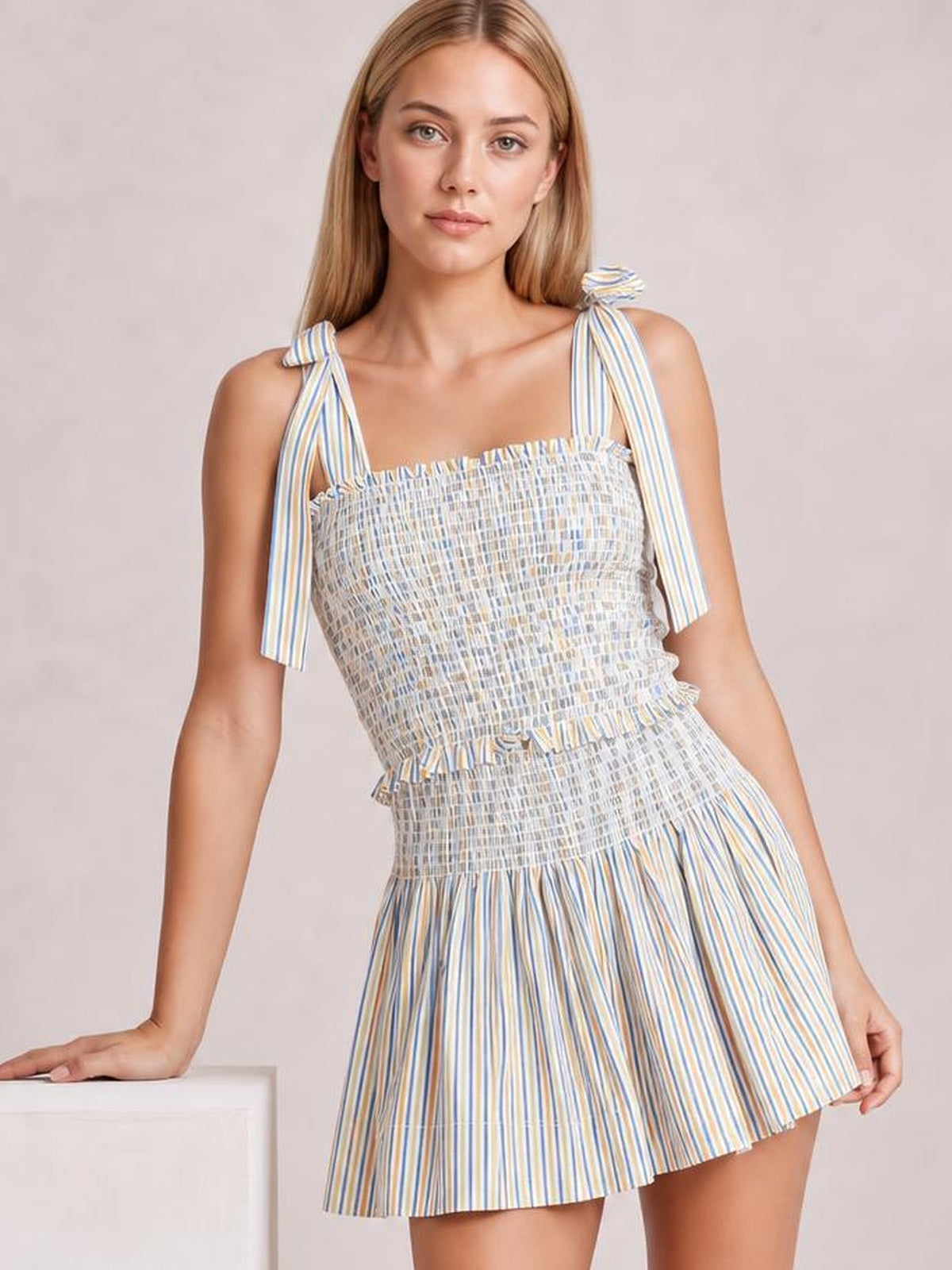 A model leans with one hand on a pedestal, wearing a blue, yellow, and white striped smocked cami and shorts set with tied shoulders and a relaxed silhouette.