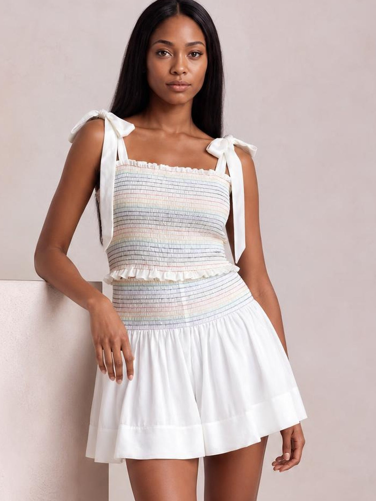 A model stands in a smocked tie-shoulder cami and shorts set with a pastel-striped bodice, white high-waisted flared shorts, and a ruffled cami hem.