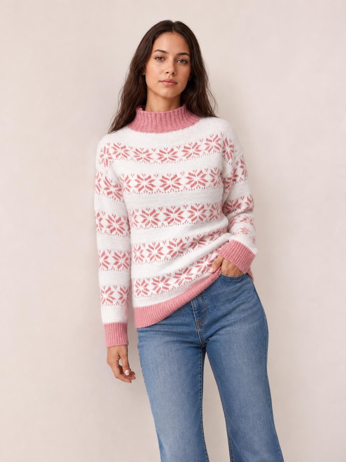 Model wears a white snowflake pattern mock neck sweater with pink ribbed collar, cuffs, and hem in a relaxed silhouette, posing with one hand in her jeans pocket.