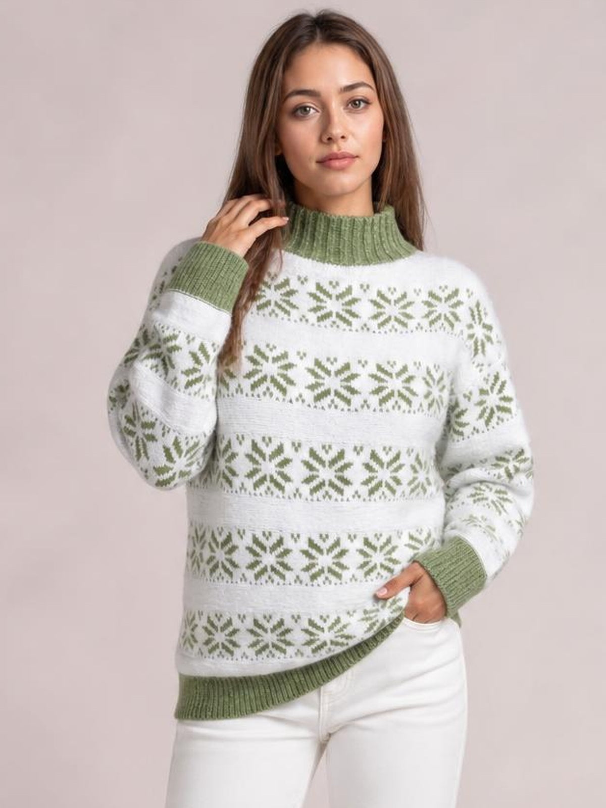 Model wears a white snowflake pattern mock neck sweater with green ribbed trim, long sleeves, and a relaxed silhouette, touching her hair and resting a hand in her pocket.