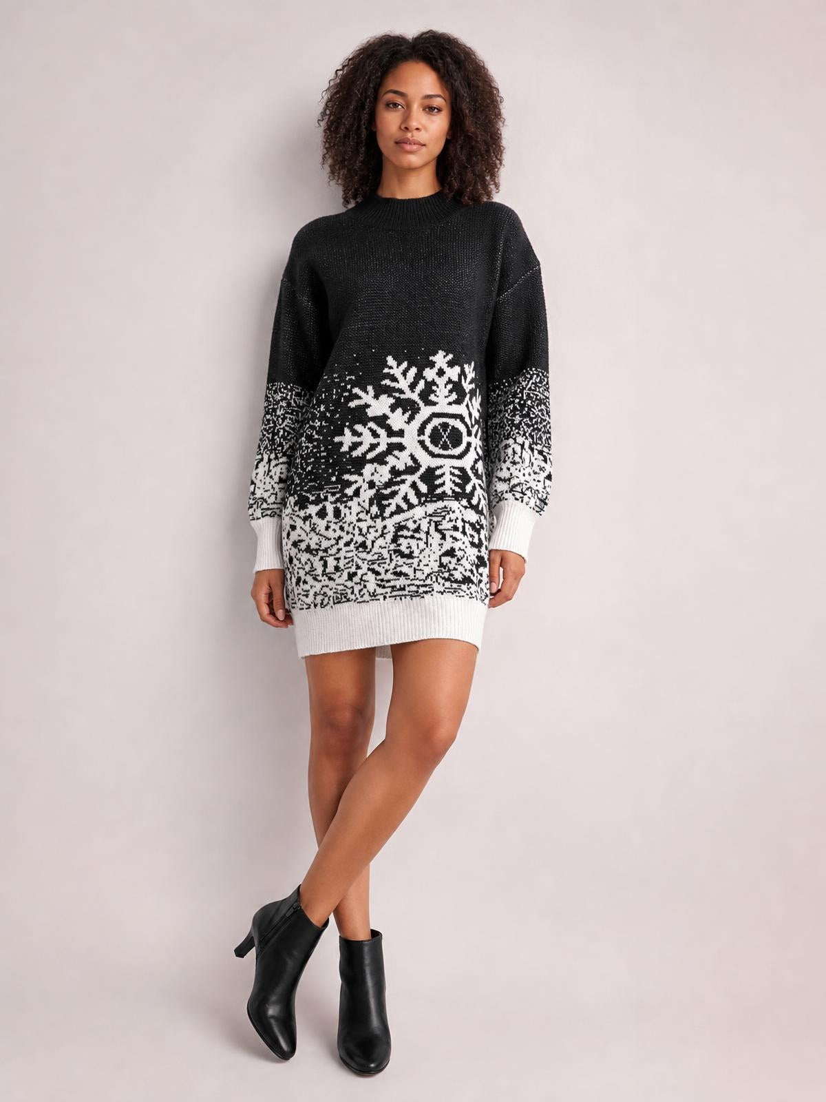 Model poses with crossed legs in a black-and-white snowflake pattern sweater dress featuring long sleeves, ribbed cuffs and hem, and a relaxed above-knee silhouette.