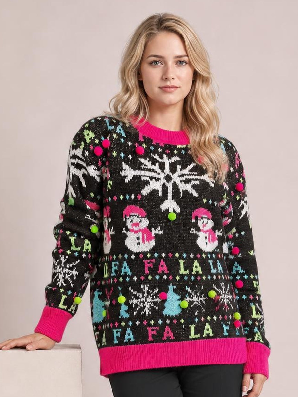A model poses in a relaxed black snowman sweater with a round neck, bright pink contrast ribbed trim, long sleeves, snowflakes, and colorful festive patterns.