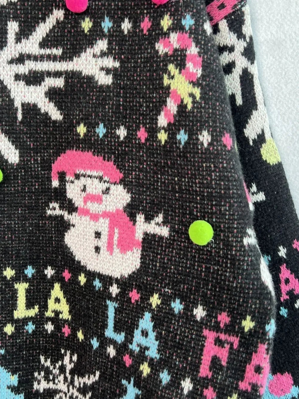 Close-up of a black knit sweater featuring a pink-and-white snowman motif, colorful lettering, and contrasting patterned trim.