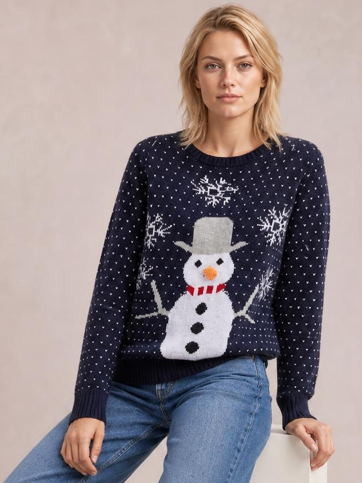 A seated model wears a navy snowman graphic raglan sleeve sweater with a relaxed silhouette, crew neckline, ribbed cuffs and hem, and festive white detailing.