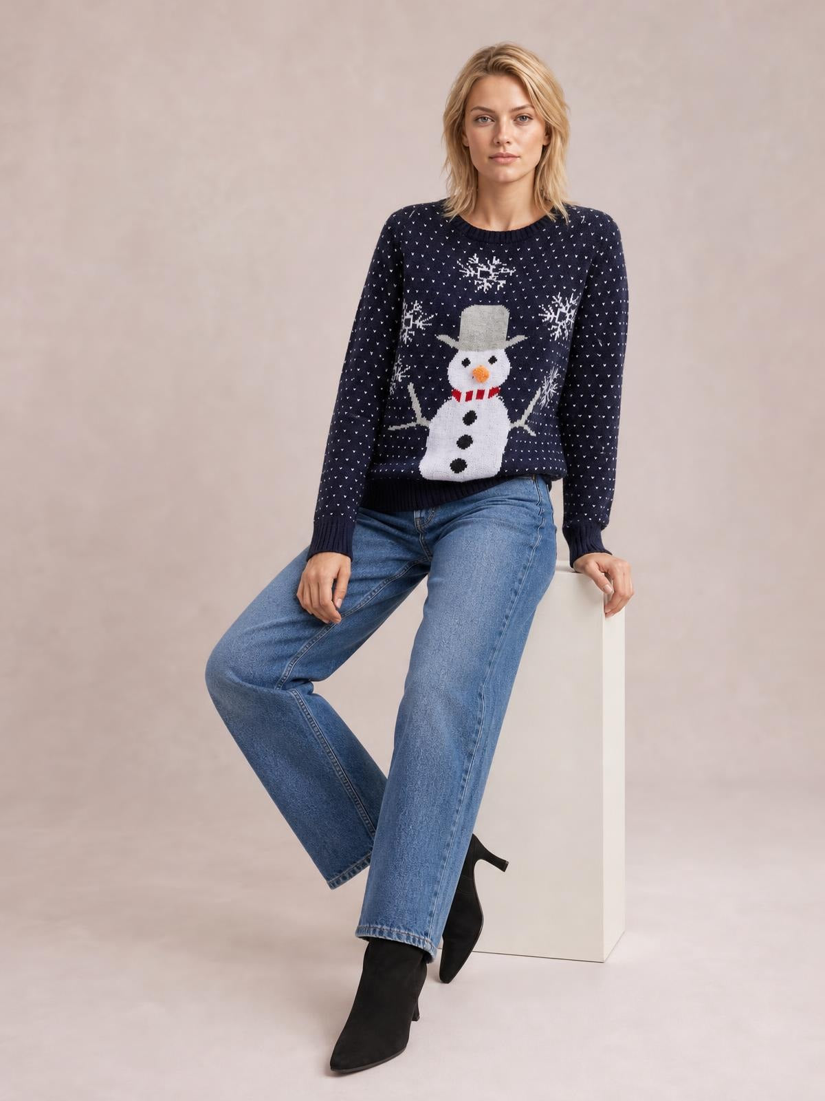 Model seated in a relaxed pose wears a navy Snowman Graphic Raglan Sleeve Sweater with a ribbed crew neck, cuffs, hem, and festive snowman design.