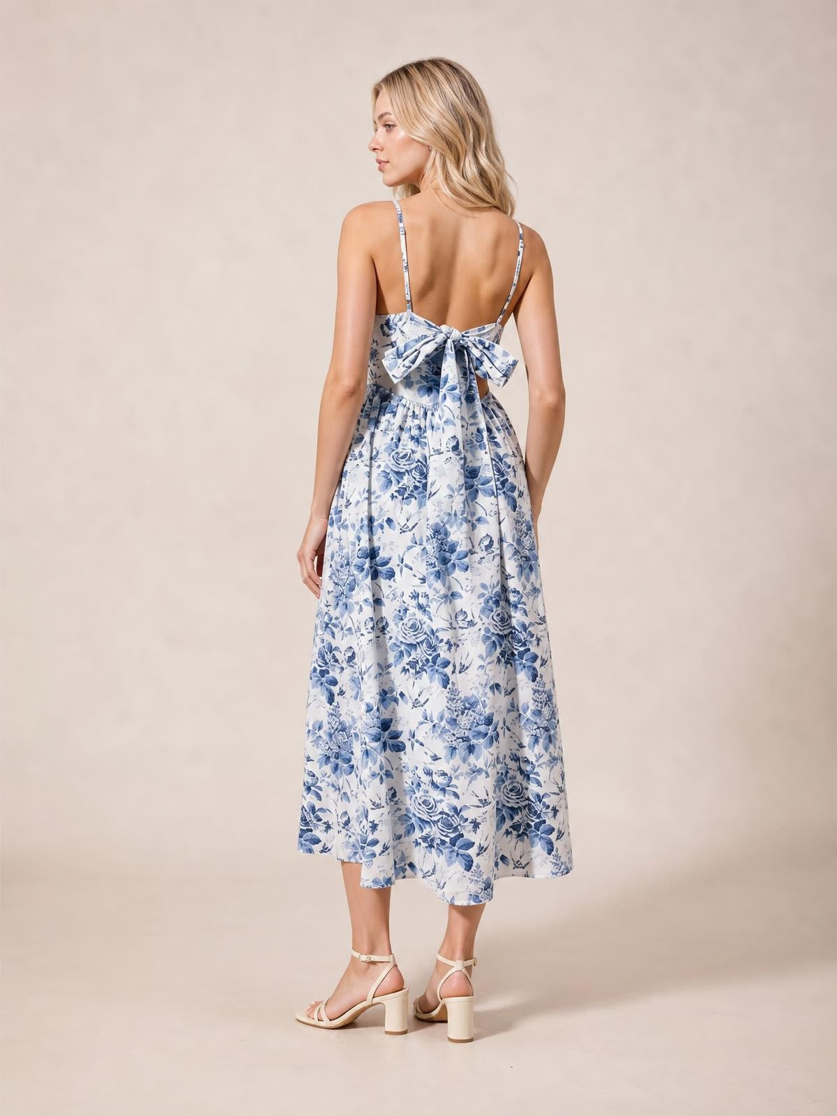 Model shown from behind in a blue-and-white floral woven dress with thin straps, a large back ribbon tie, gathered waist, and flowing midi silhouette.