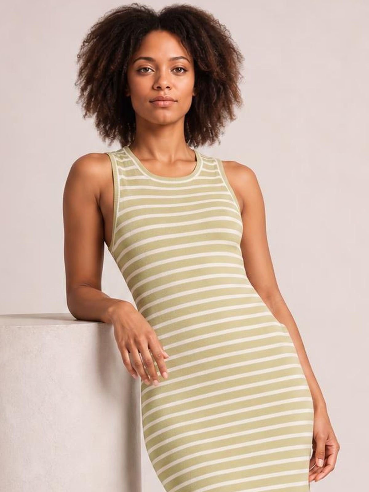 A model leans against a ledge wearing a sage green and white striped sleeveless sweater knit dress with a scoop neck and fitted silhouette.