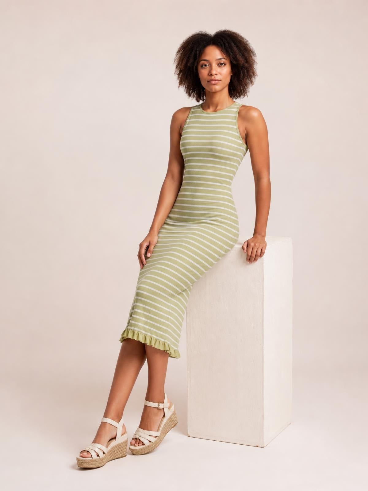 Model poses in a fitted olive green and white striped sleeveless sweater-knit maxi dress with a ruffled hem and softly curved silhouette.