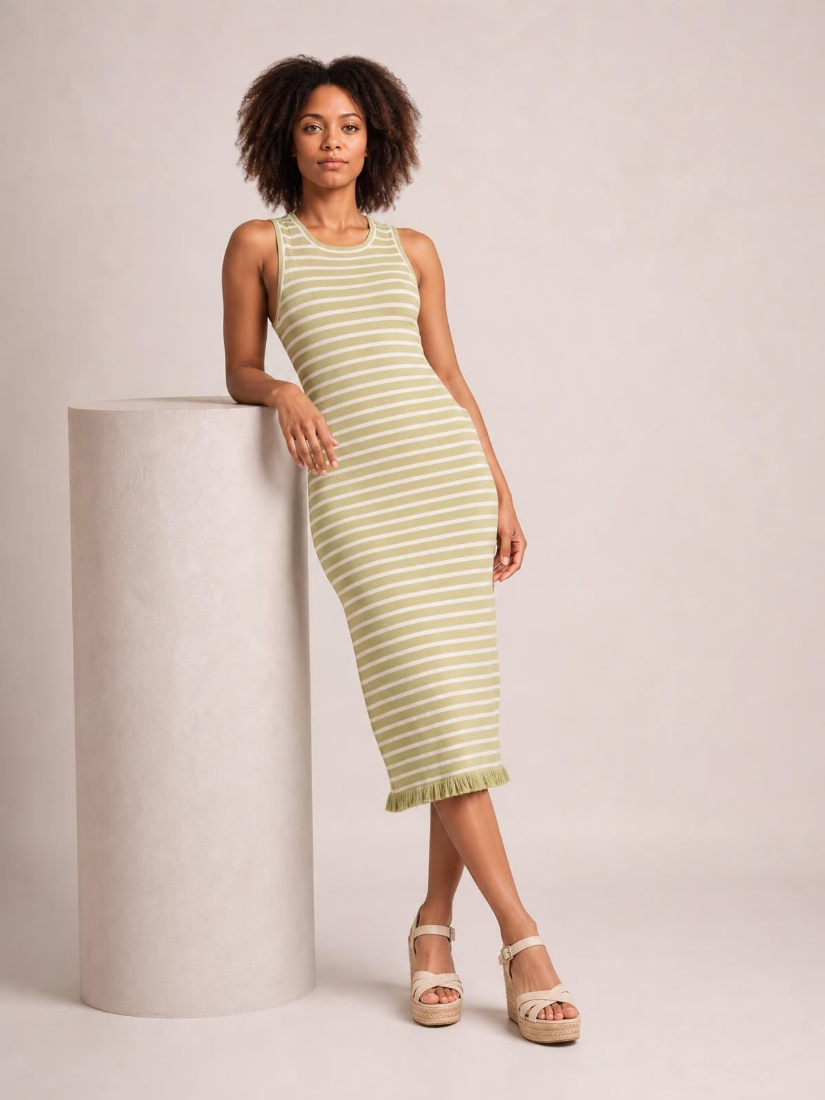 A model leans in a fitted olive and white striped sleeveless sweater-knit maxi dress with a scoop neckline, streamlined silhouette, and ruffled hem.