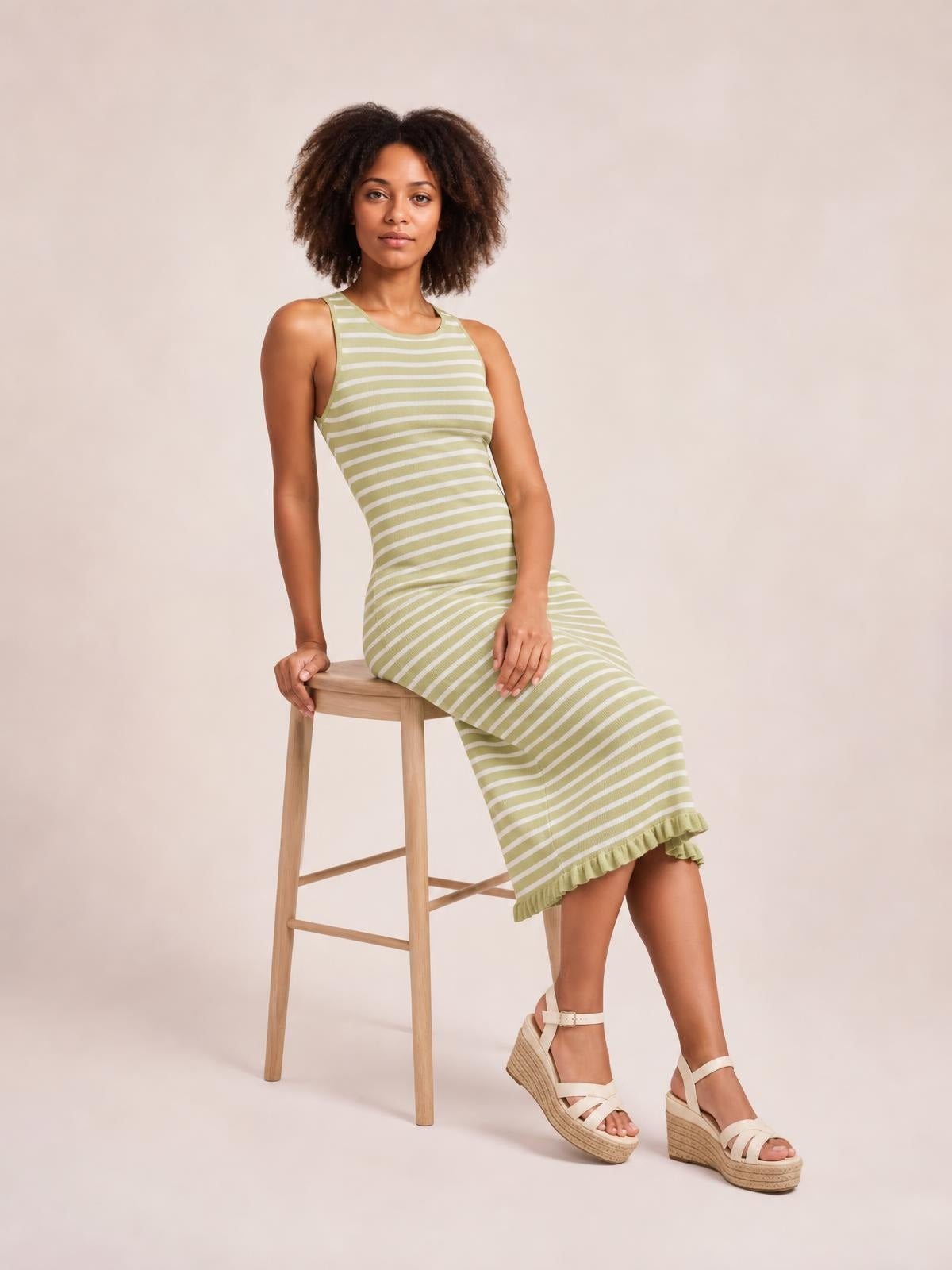 Model seated on a stool wears a sage green and white striped sleeveless sweater-knit maxi dress with a fitted silhouette and ruffled hem.
