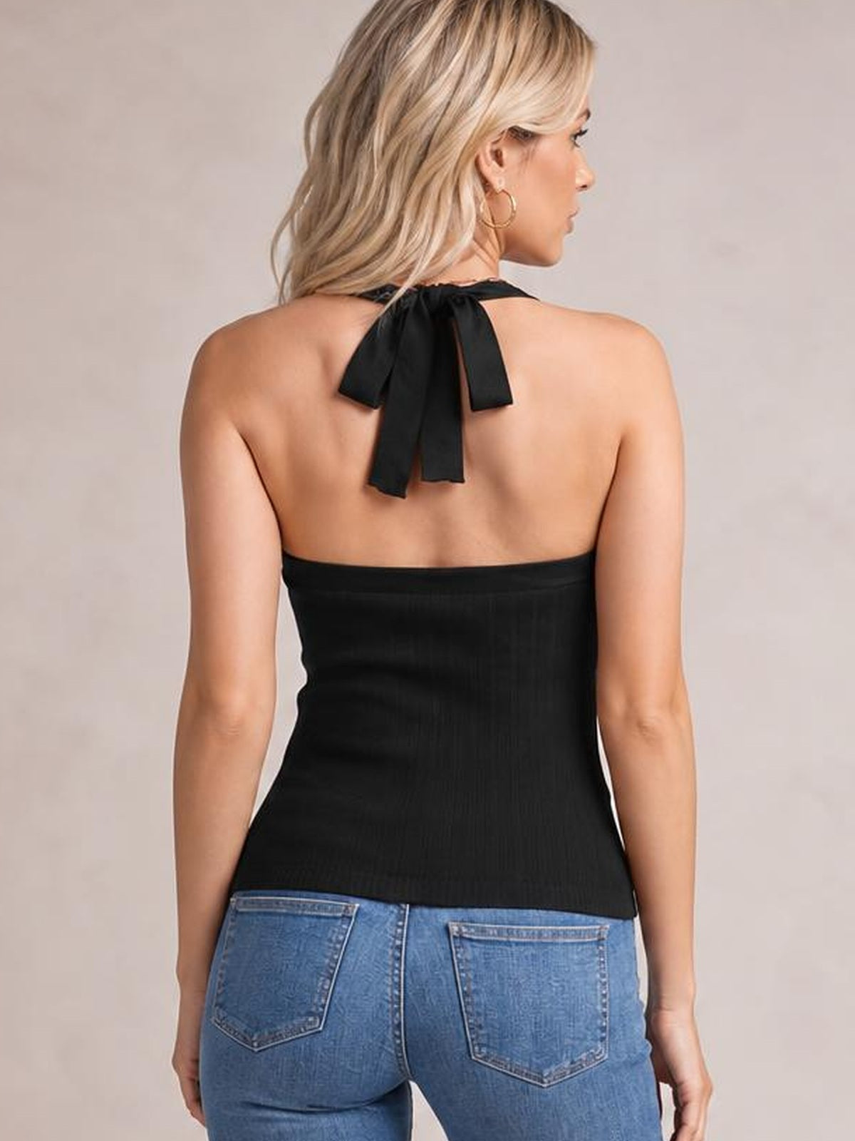 A model faces away in a fitted black halter neck knit cami with a tied back neckline, subtle vertical texture, and a straight hem over blue jeans.