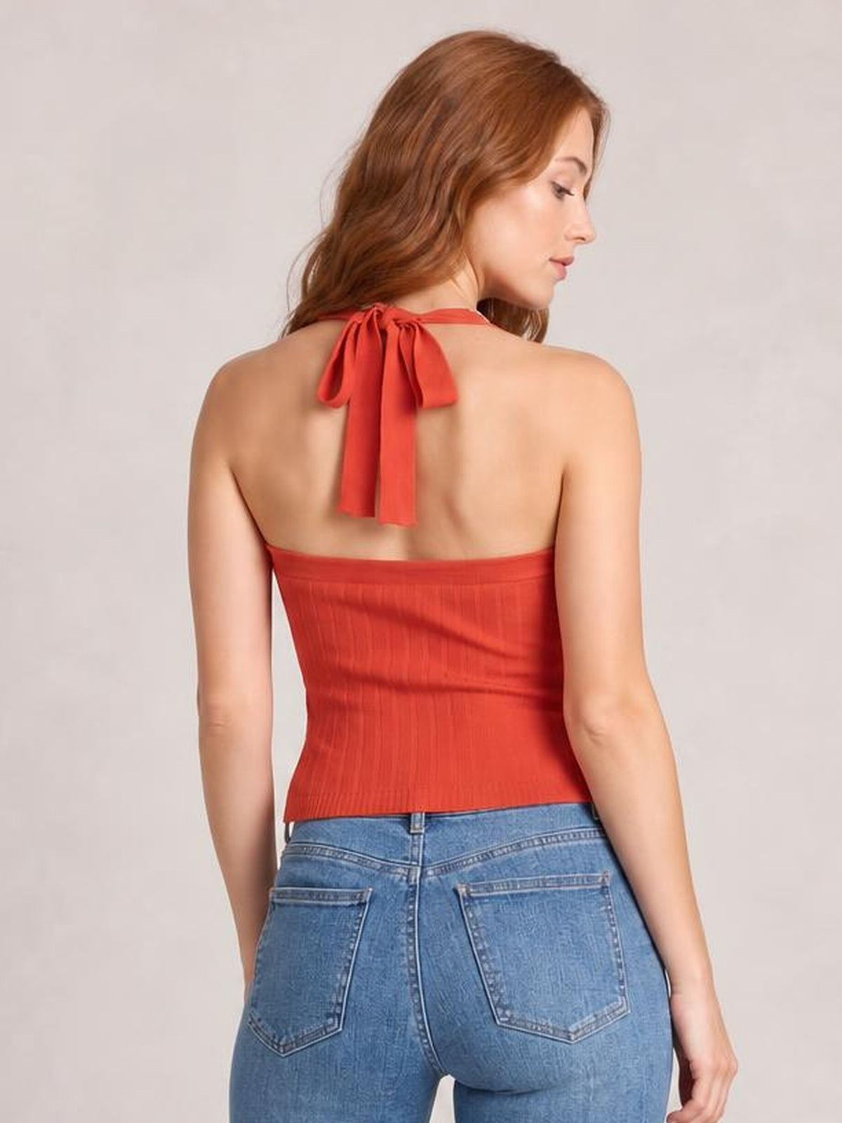 A model faces away wearing a solid red halter neck knit cami with a fitted silhouette, open back, and long tied neck straps.