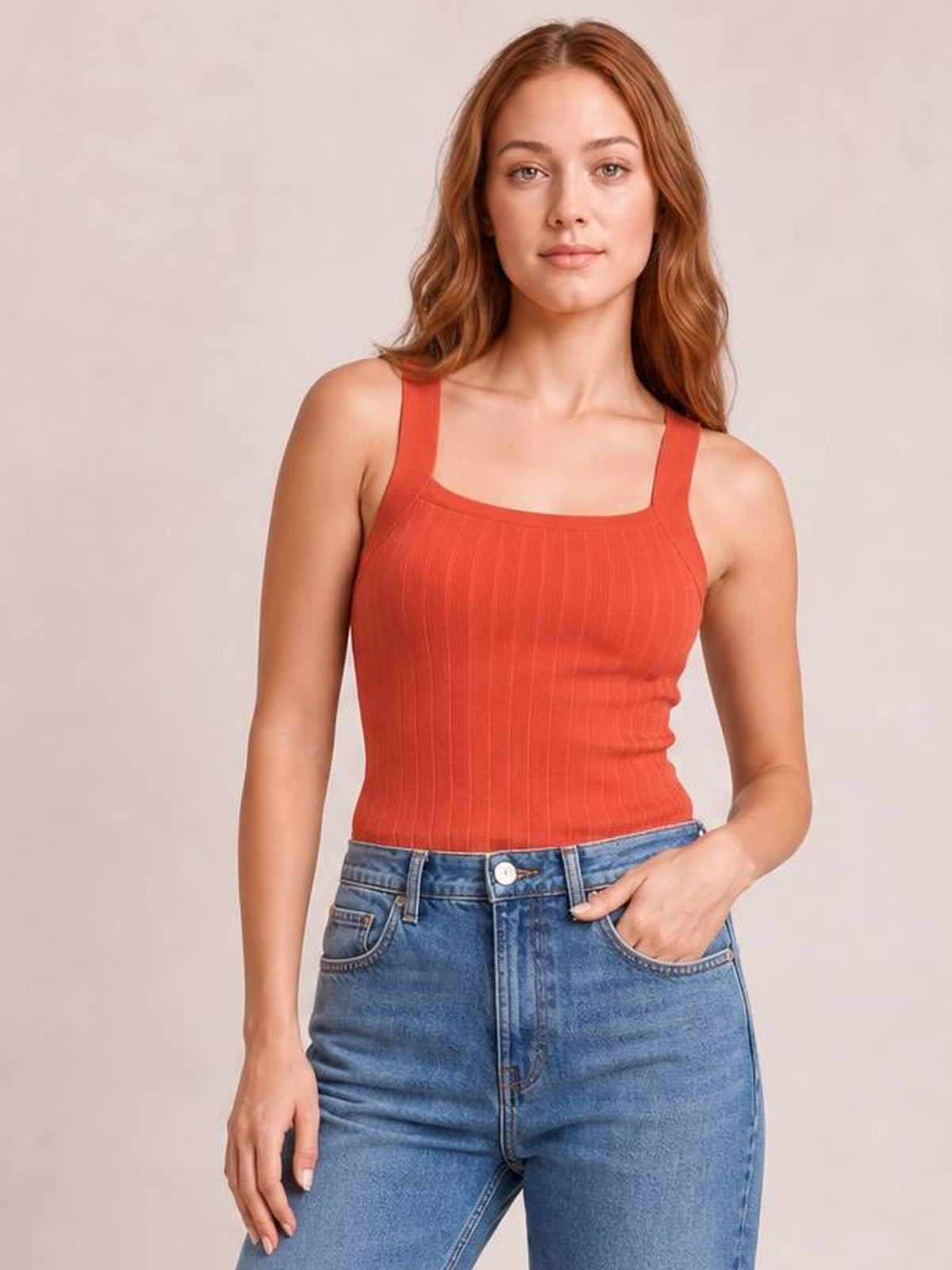 Model wears a solid coral knit cami with a square neckline, wide shoulder straps, subtle vertical texture, and a fitted silhouette with one hand in her jeans pocket.