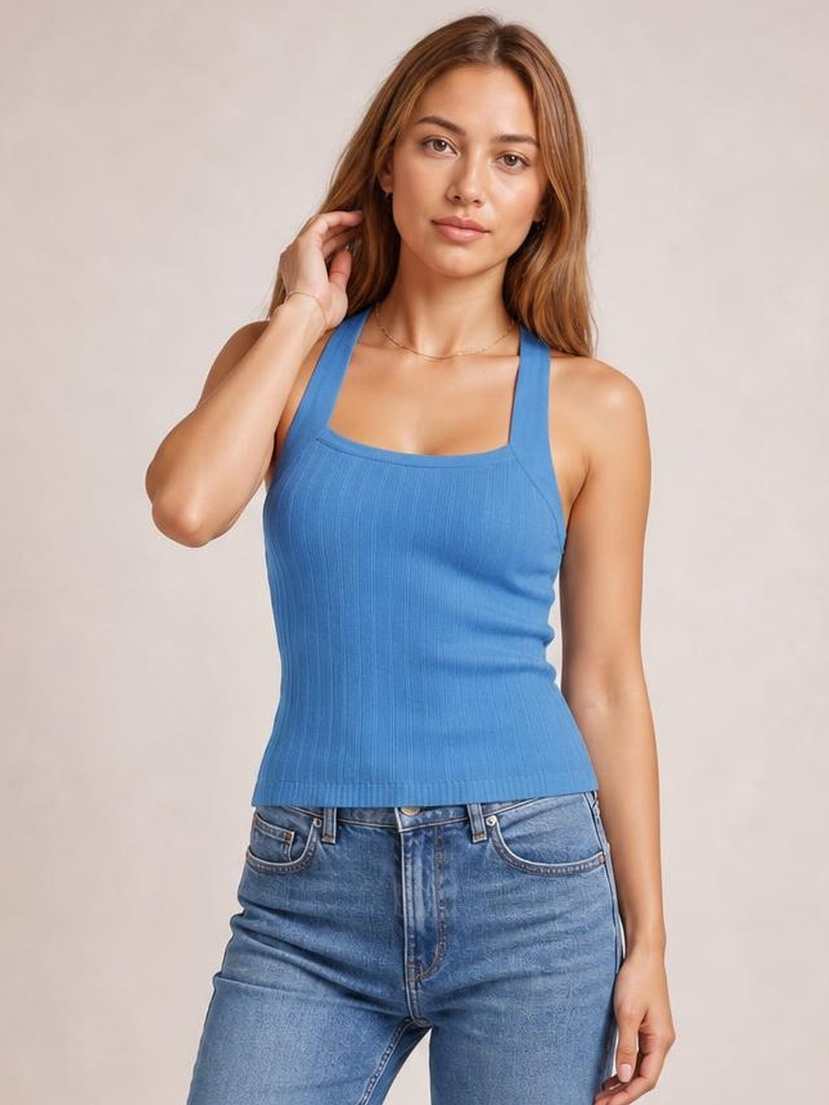 A model wears a solid blue halter neck knit cami with wide straps, square neckline, ribbed texture, fitted silhouette, and subtle stretch while touching her hair.