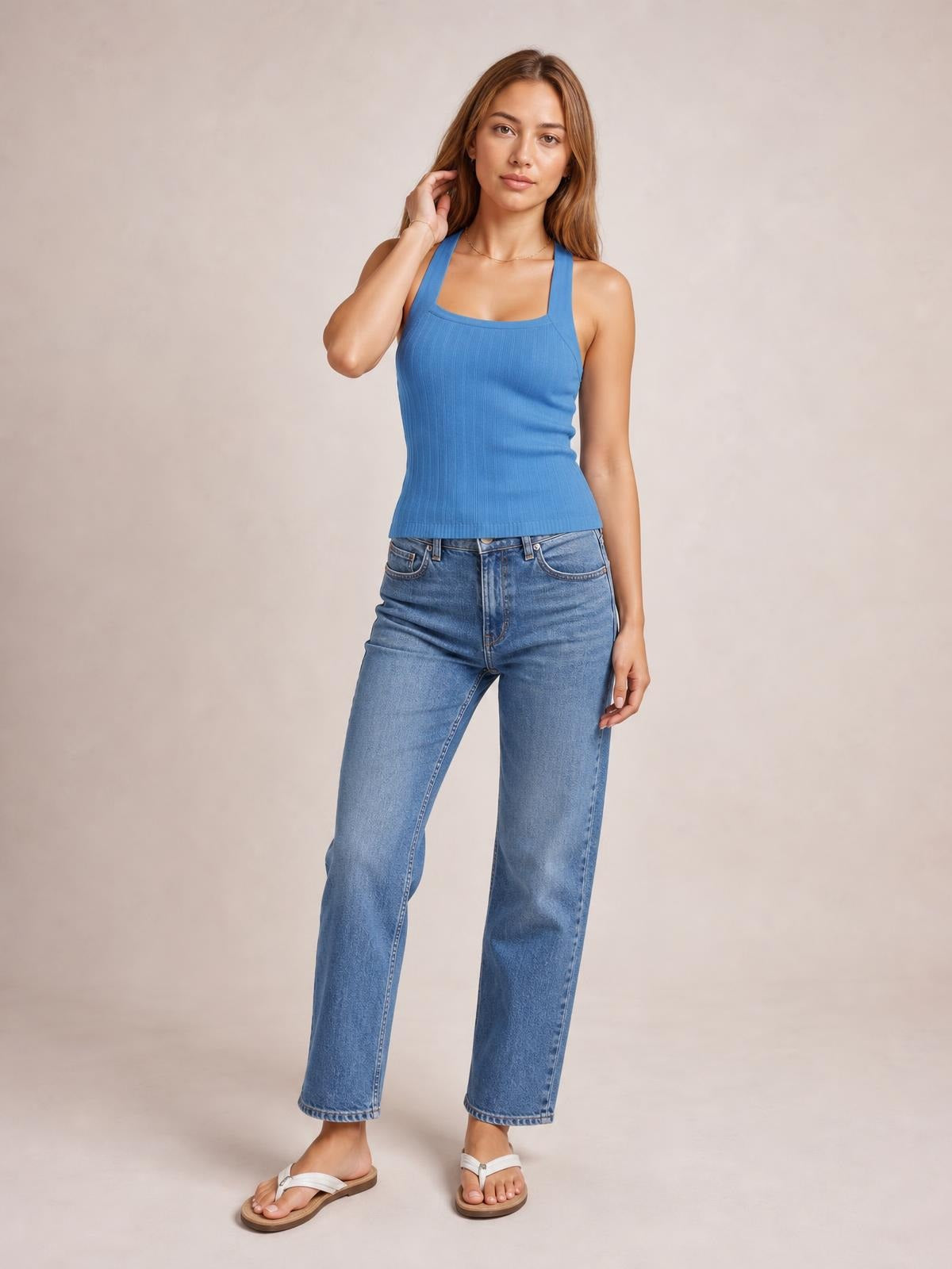 A model poses in a solid blue knit cami with wide straps, a square neckline, ribbed construction, and a fitted silhouette, paired with jeans.