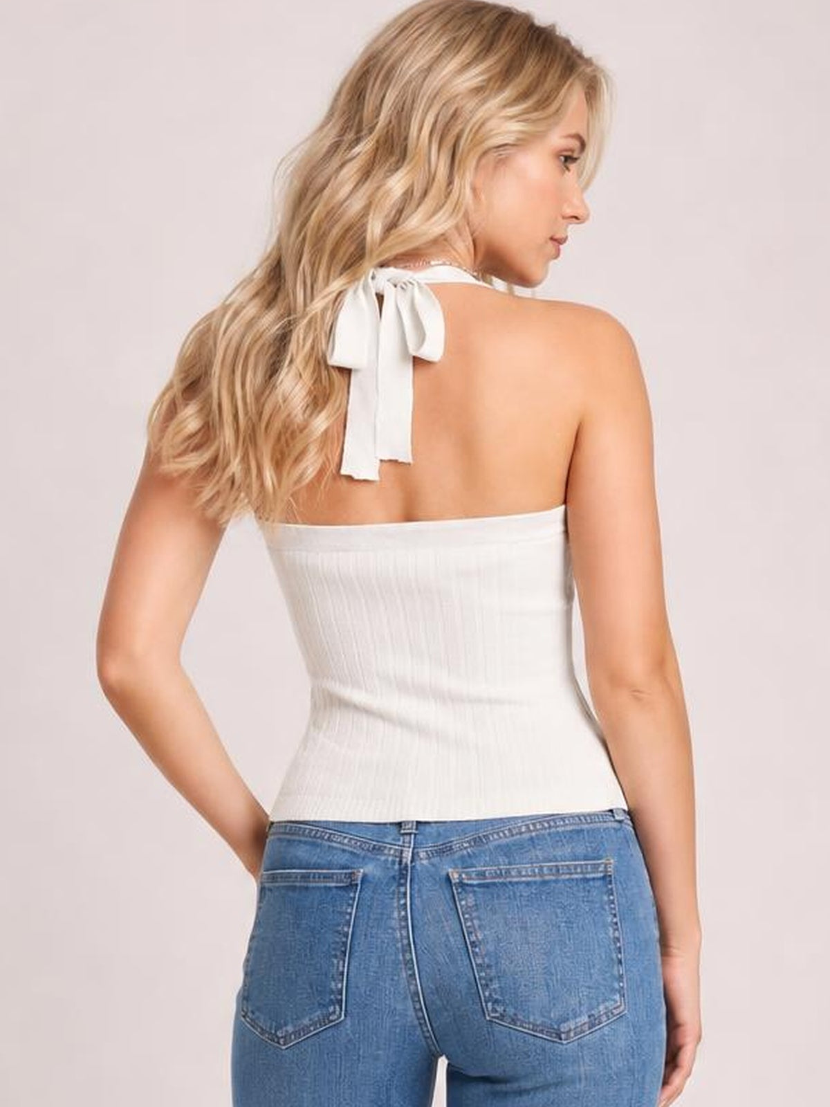 Back view of a model wearing a white halter neck knit cami with a tied neck bow, ribbed texture, fitted silhouette, and one hand resting at her hip.