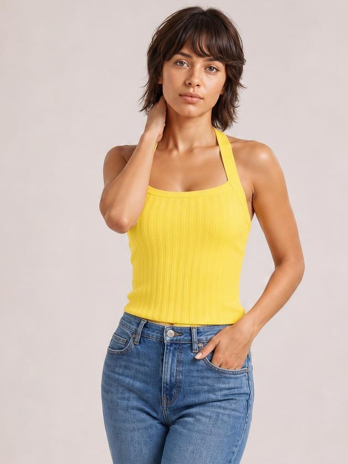 Model wearing a solid yellow halter neck knit cami with ribbed texture, fitted silhouette, and wide straps, posing with one hand at her neck and the other in her jeans pocket.