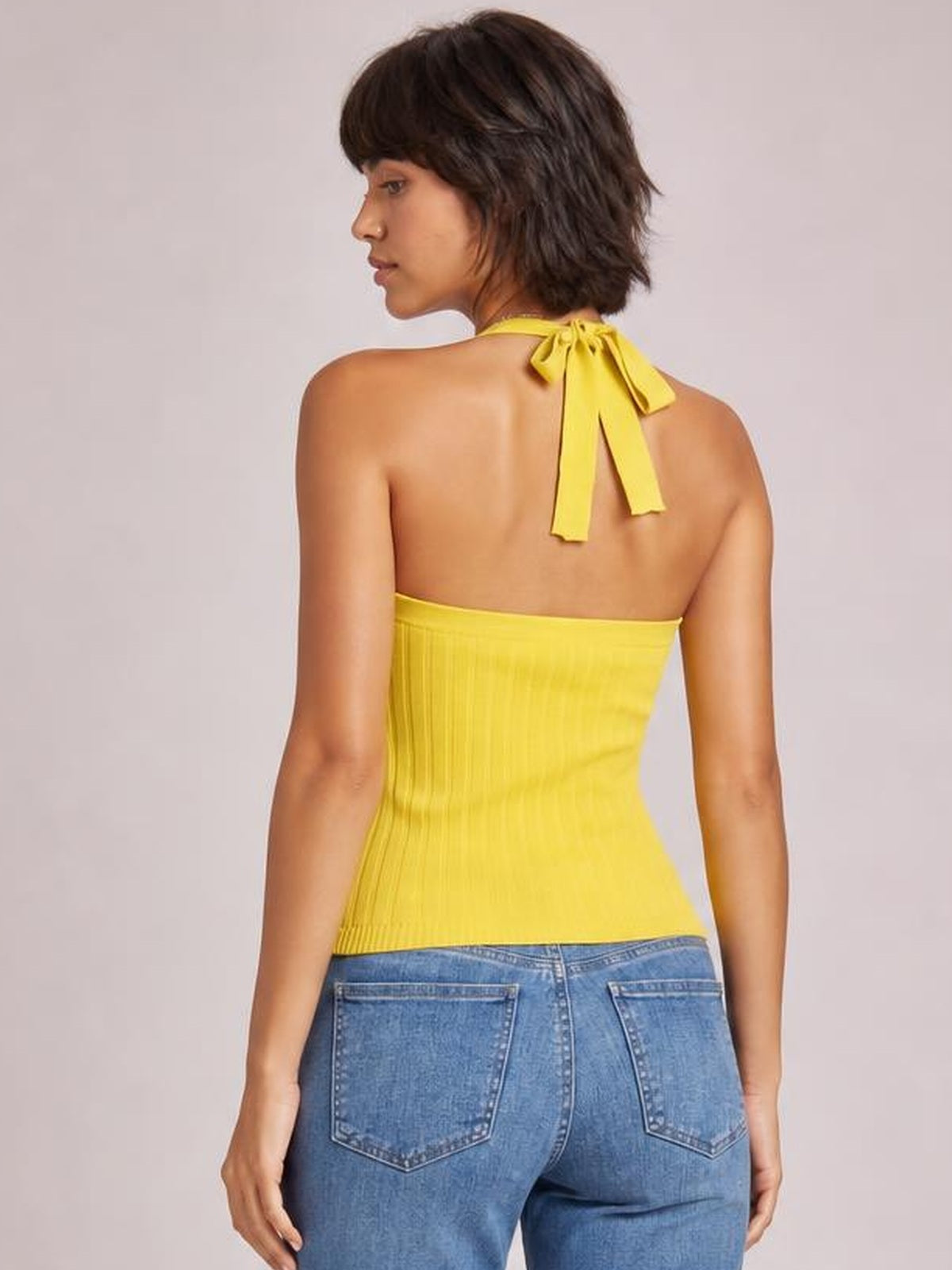 A model looks over her shoulder wearing a solid yellow halter neck knit cami with a tied neckline, ribbed texture, and fitted silhouette.