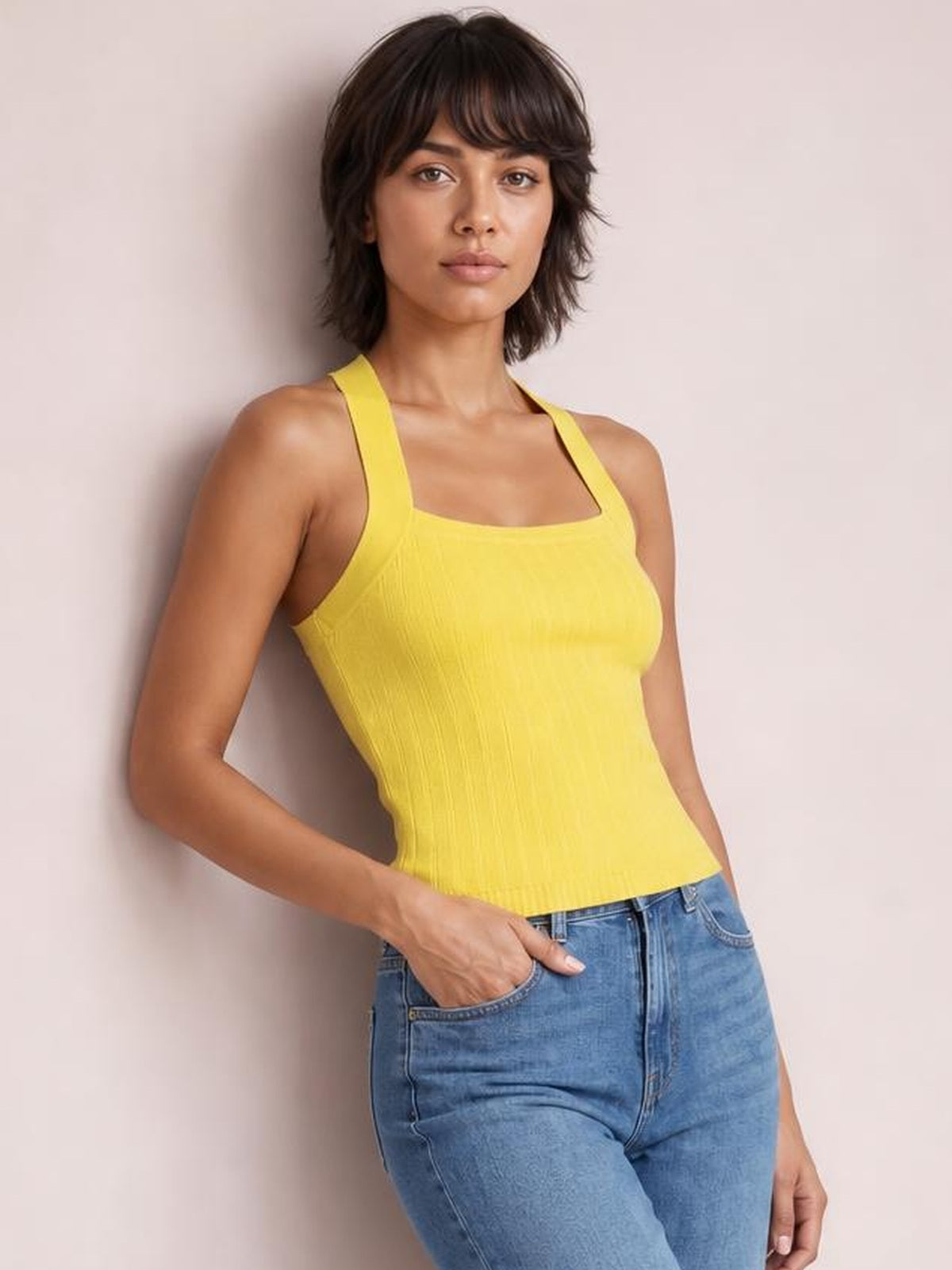 Model wears a bright yellow solid-color knit cami with wide shoulder straps, a square neckline, fitted silhouette, and subtle vertical texture while leaning with one hand in jeans pocket.