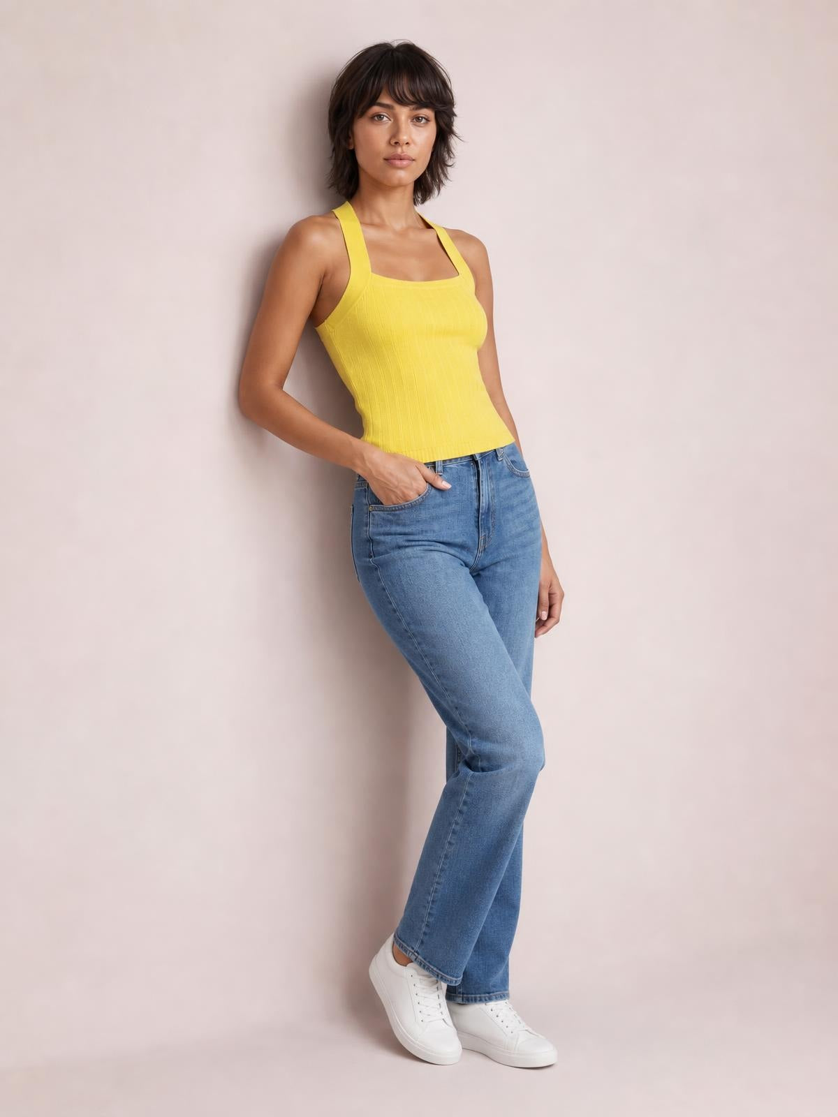 A model wears a solid yellow knit cami with wide straps, a square neckline, ribbed texture, and fitted silhouette while leaning with one hand in her jeans pocket.