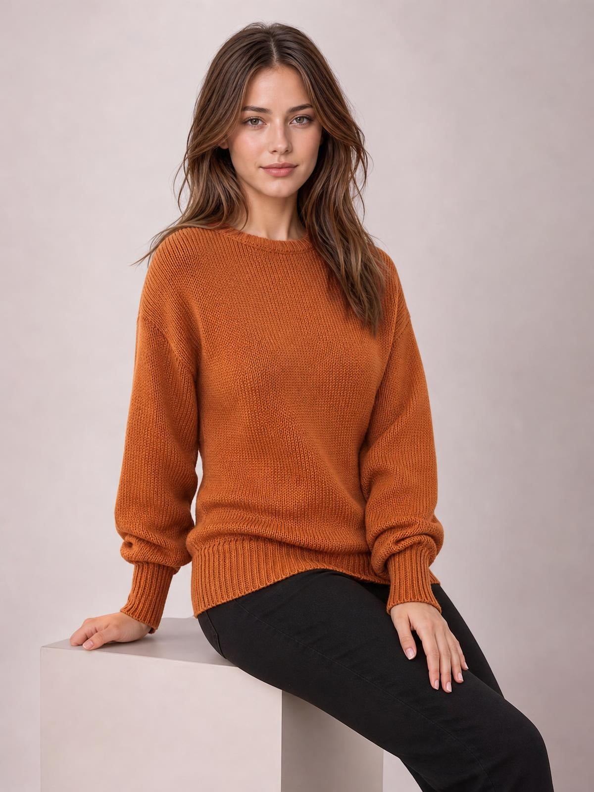 A seated model wears a solid rust-orange ribbed sweater with a relaxed drop-shoulder silhouette, crew neck, ribbed cuffs, and a ribbed hem.