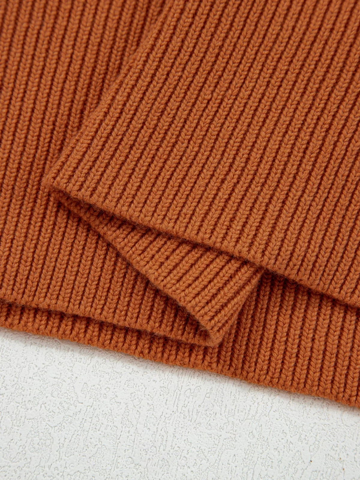 Solid rust-brown sweater close-up showing dense vertical ribbed knit texture and overlapping ribbed trim at the hem or cuff.