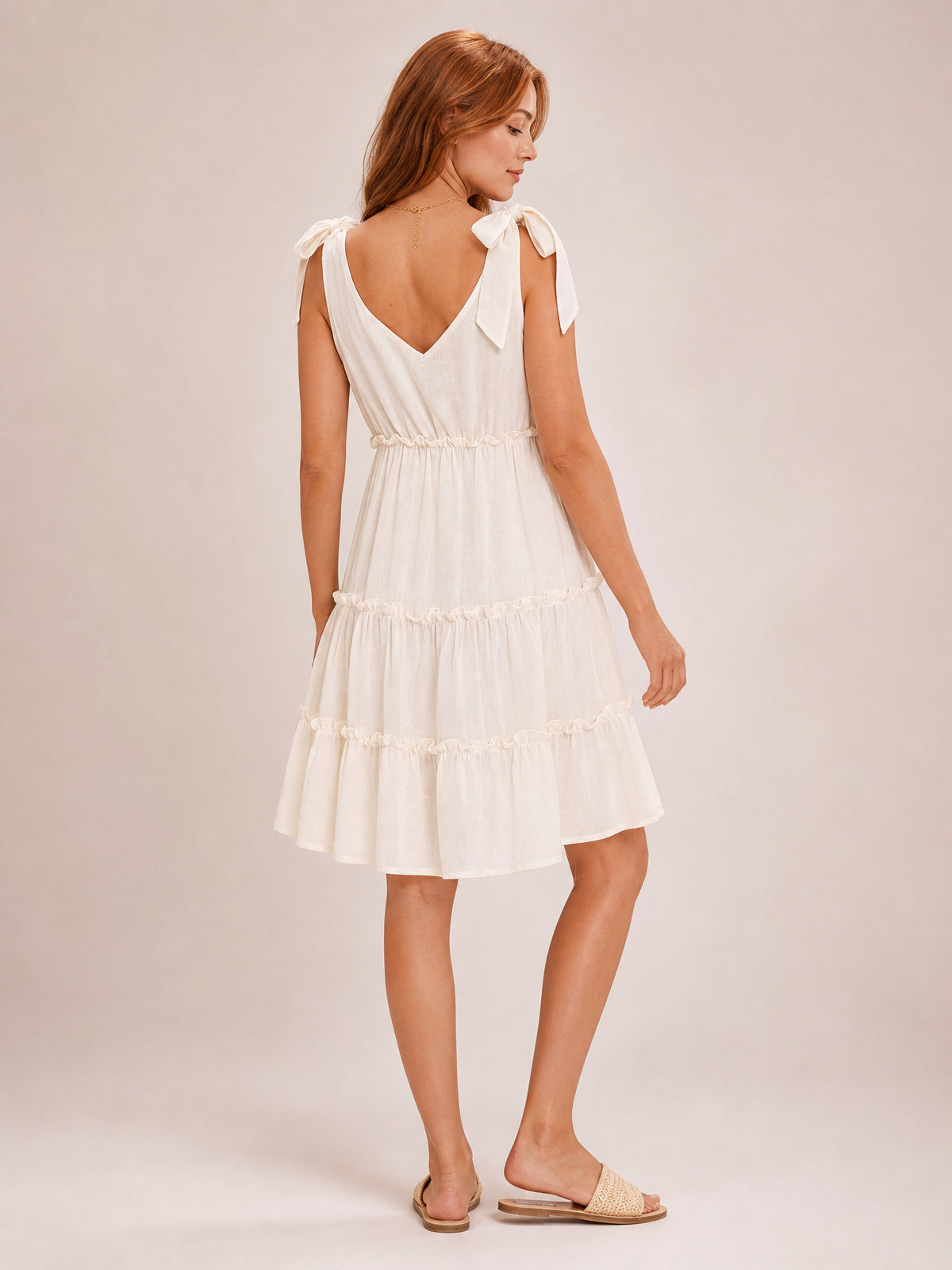 A model turns to show a cream mini dress with tied shoulder straps, a V-back, defined waist, and flowing tiered silhouette trimmed with delicate ruffles.