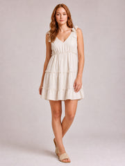 Model stands with crossed legs in a beige mini dress featuring a V-neck, tied shoulder straps, defined waist, and ruffle-trimmed tiered silhouette.