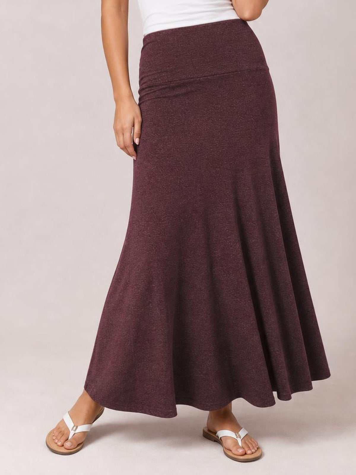 A model stands in a burgundy solid elastic waist maxi skirt with a fitted upper silhouette, flowing flare, and ankle-length hem.