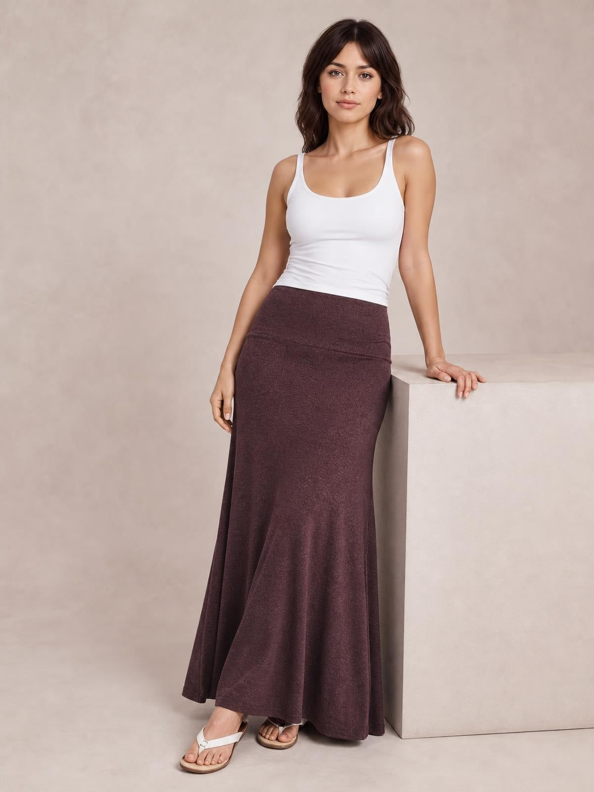 A model leans against a block wearing a solid dark brown elastic-waist maxi skirt with a smooth, fitted upper silhouette and flowing hem.