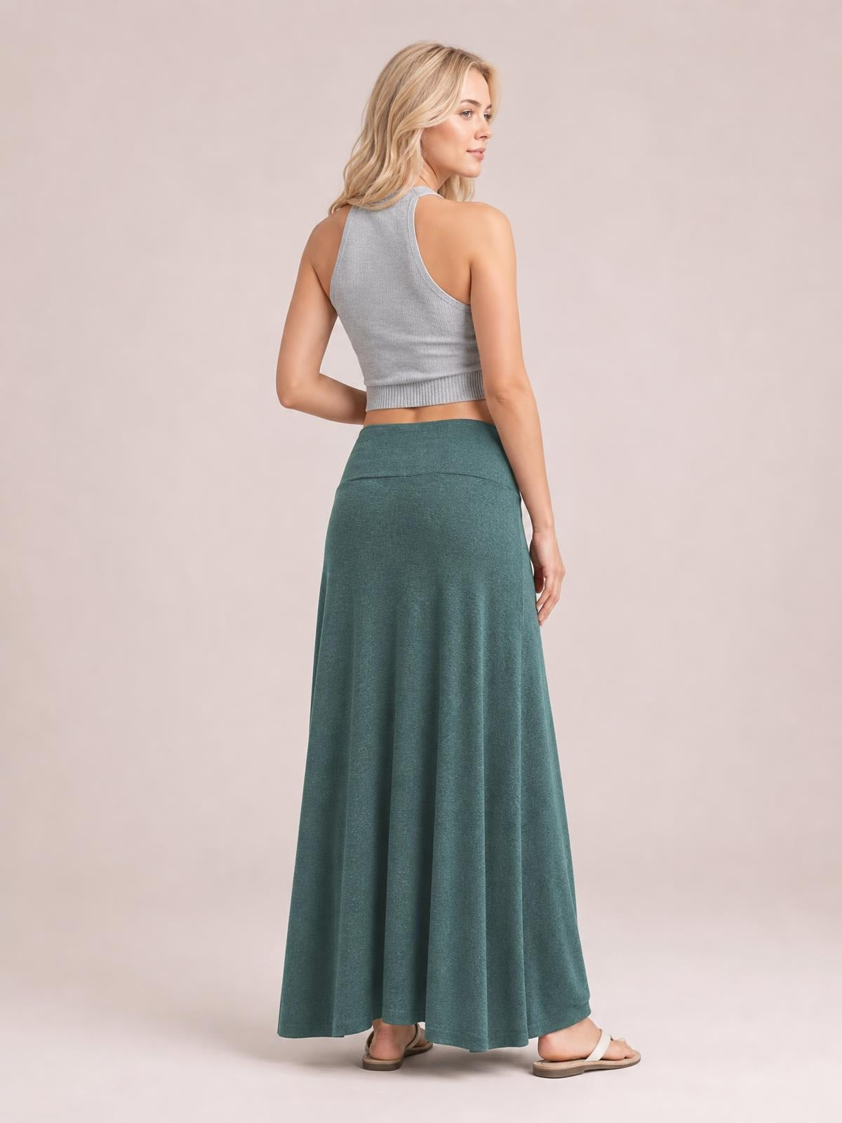 A model poses turned away in a solid teal maxi skirt with a wide elastic waist, smooth construction, and a long, flowy silhouette that drapes to the ankles.