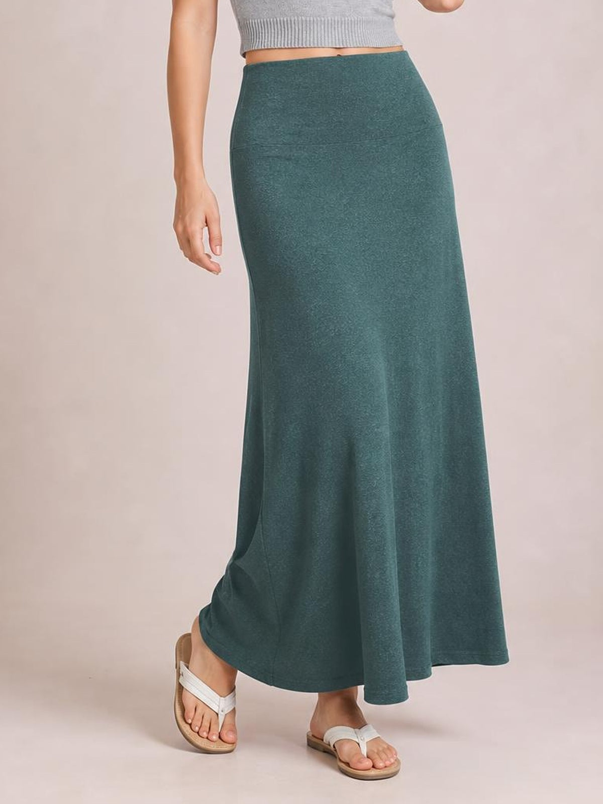 A model wears a solid teal elastic-waist maxi skirt with a fitted silhouette through the hips, flowing toward the ankle-length hem as she poses in sandals.