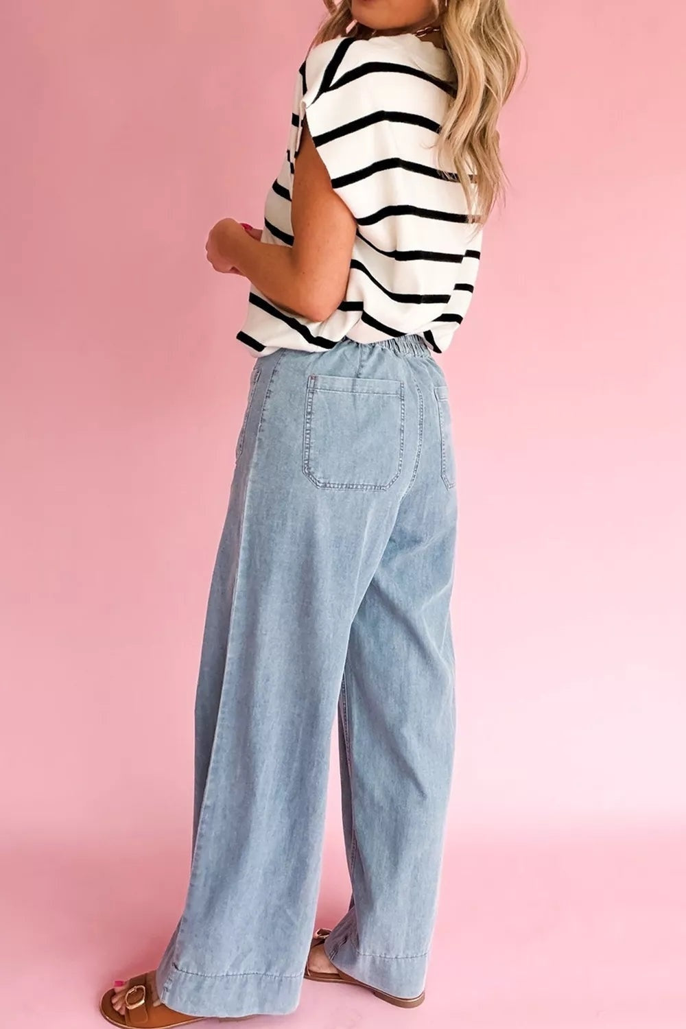 A model poses turned away in light blue full-length jeans with a relaxed wide-leg silhouette, gathered waistband, and visible back patch pocket.