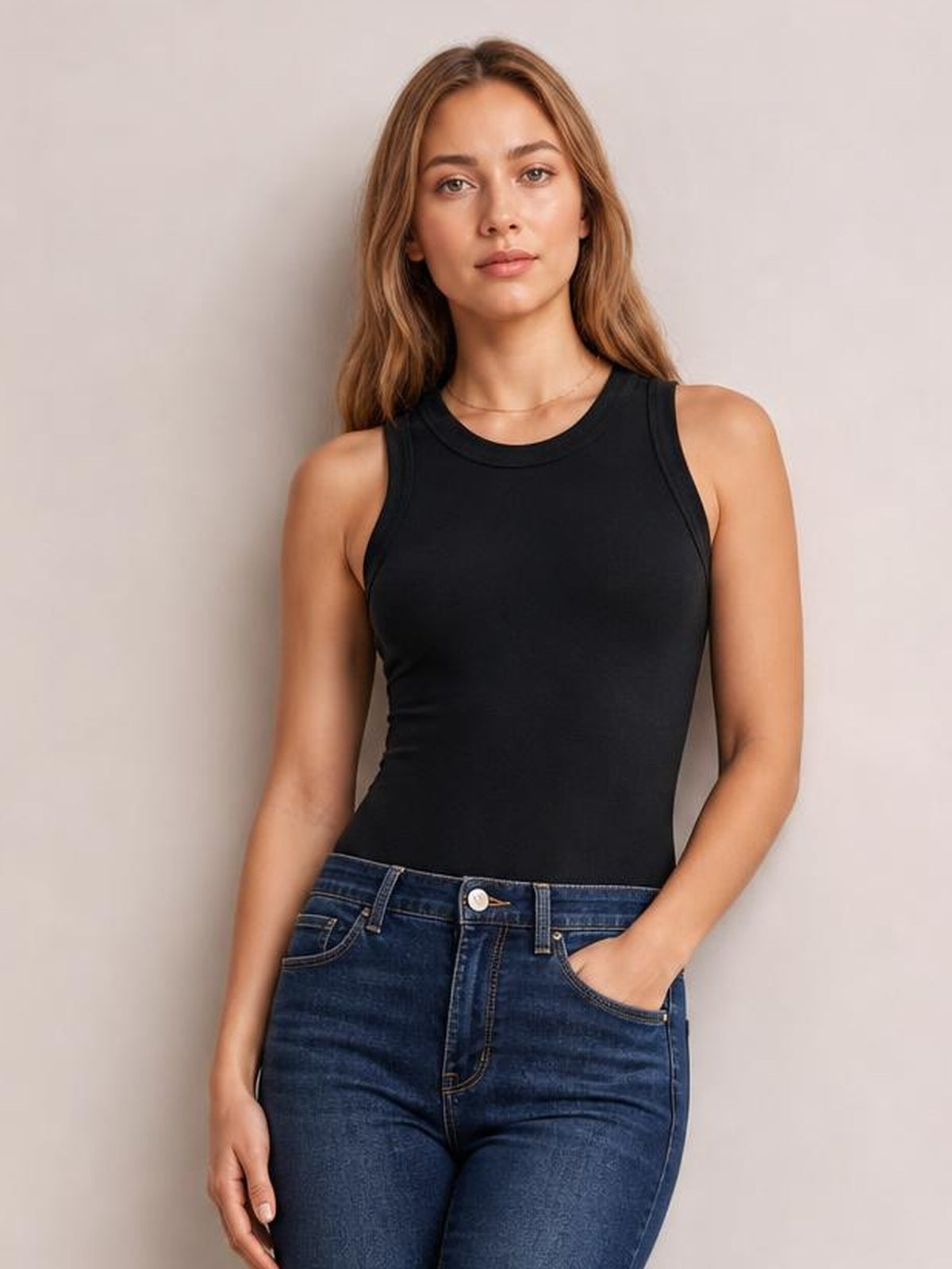 Model wears a solid black round neck tank with wide shoulder straps and a fitted silhouette, standing with one hand in her jeans pocket.