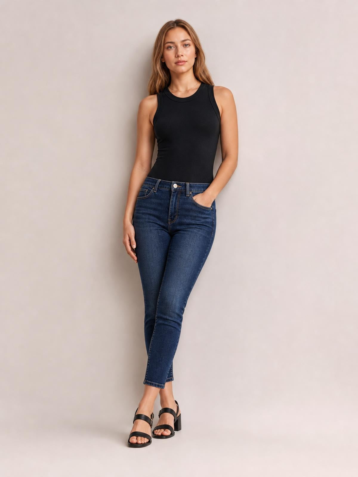 Model wears a solid black round neck tank with a fitted sleeveless silhouette and slight stretch, paired with cropped blue jeans while standing with one hand in a pocket.