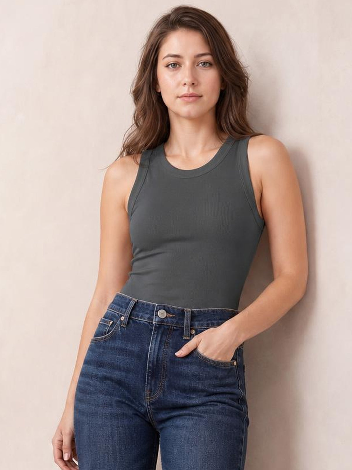 A model wears a solid charcoal round neck tank with wide shoulder straps, bound edges, and a fitted sleeveless silhouette, posing with one hand in her jeans pocket.