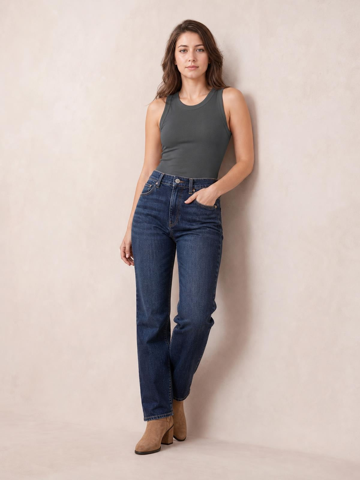 A model wears a solid charcoal round-neck tank with a sleeveless cut, fitted silhouette, and clean seamed edges, standing with one hand in her jeans pocket.