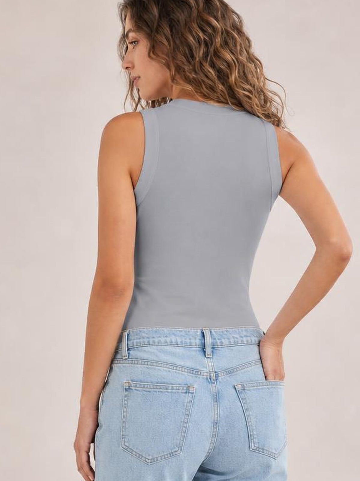 Back view of a woman wearing a solid light blue-gray round neck tank with a fitted sleeveless silhouette, posed with one hand on her hip.