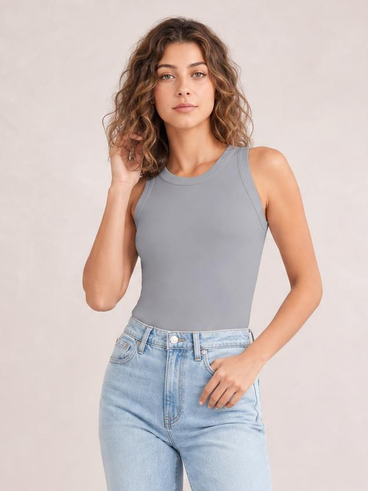 Model wears a solid gray round neck tank with wide shoulder straps and a fitted silhouette, posing with one hand in her jeans pocket and one in her hair.