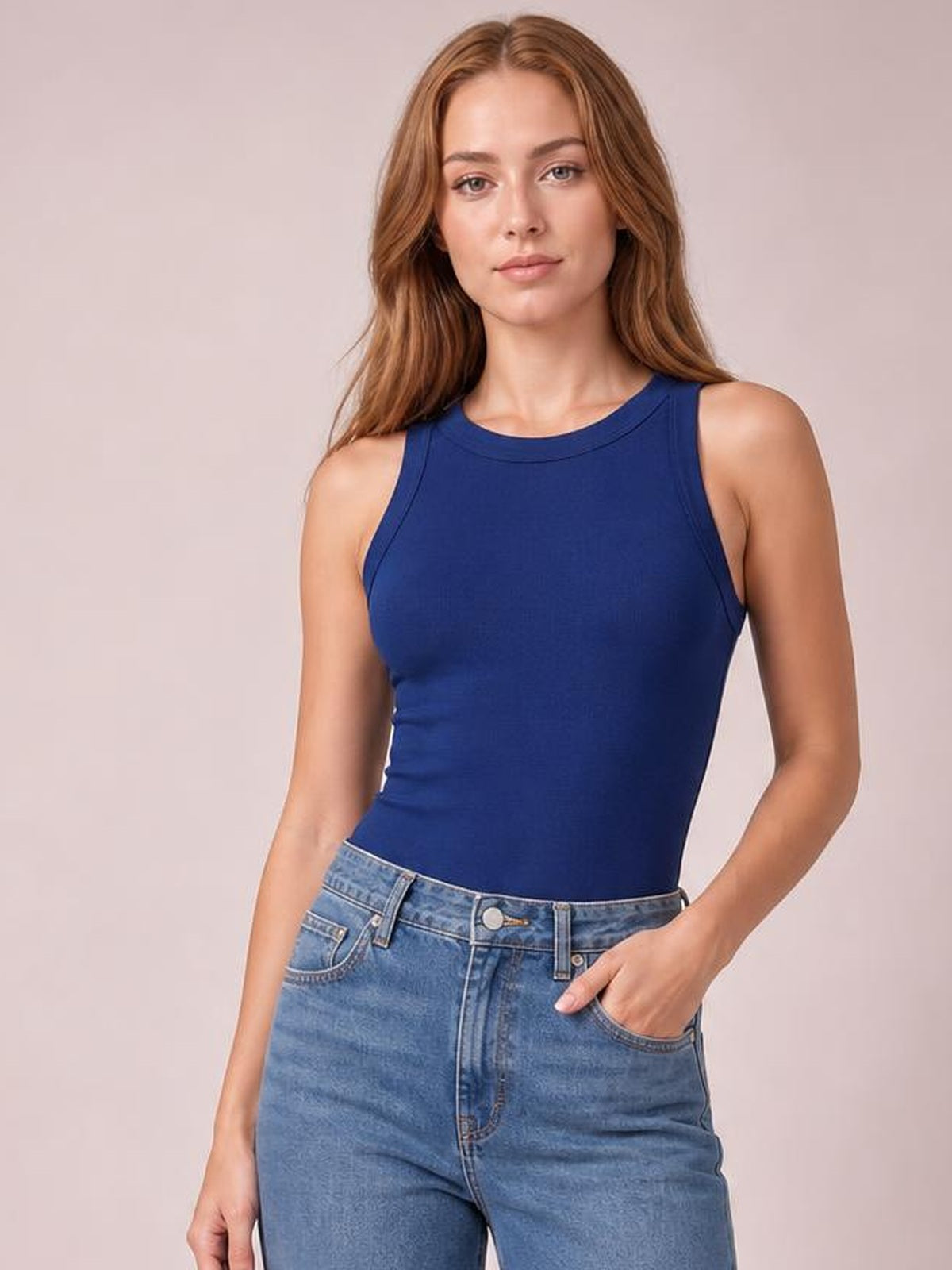 Model wears a solid navy blue tank with a round neckline, sleeveless cut, fitted silhouette, and subtle stretch, posing with one hand in her jeans pocket.