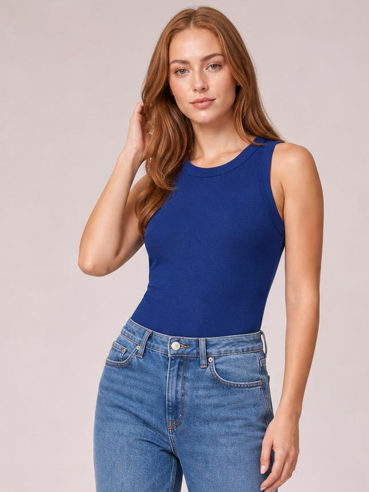 Model poses with one hand in her hair, wearing a solid blue round neck tank with a fitted sleeveless silhouette tucked into high-rise jeans.