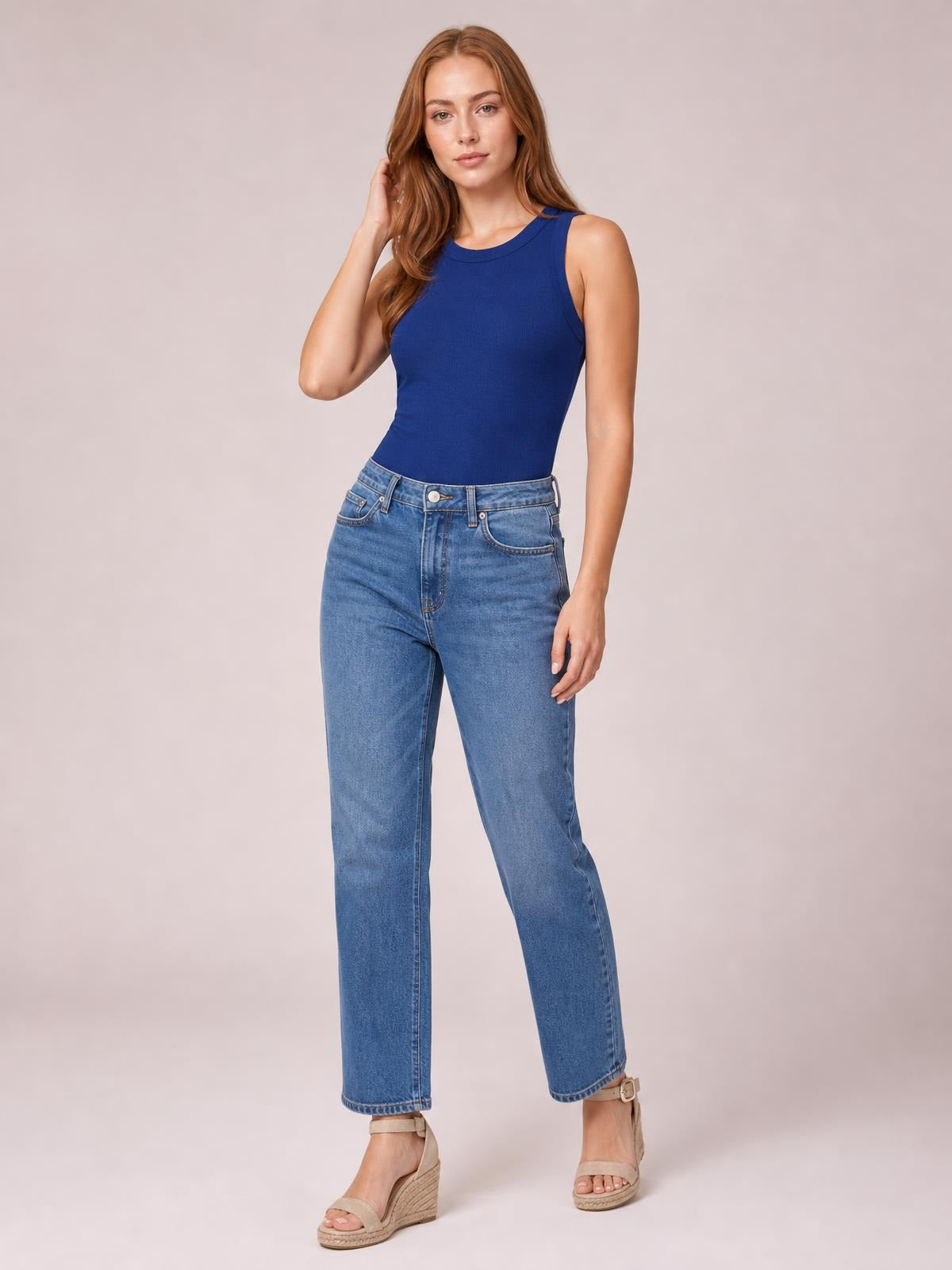 A model poses with one hand in her hair, wearing a solid cobalt blue tank with a round neck, sleeveless cut, and fitted silhouette.