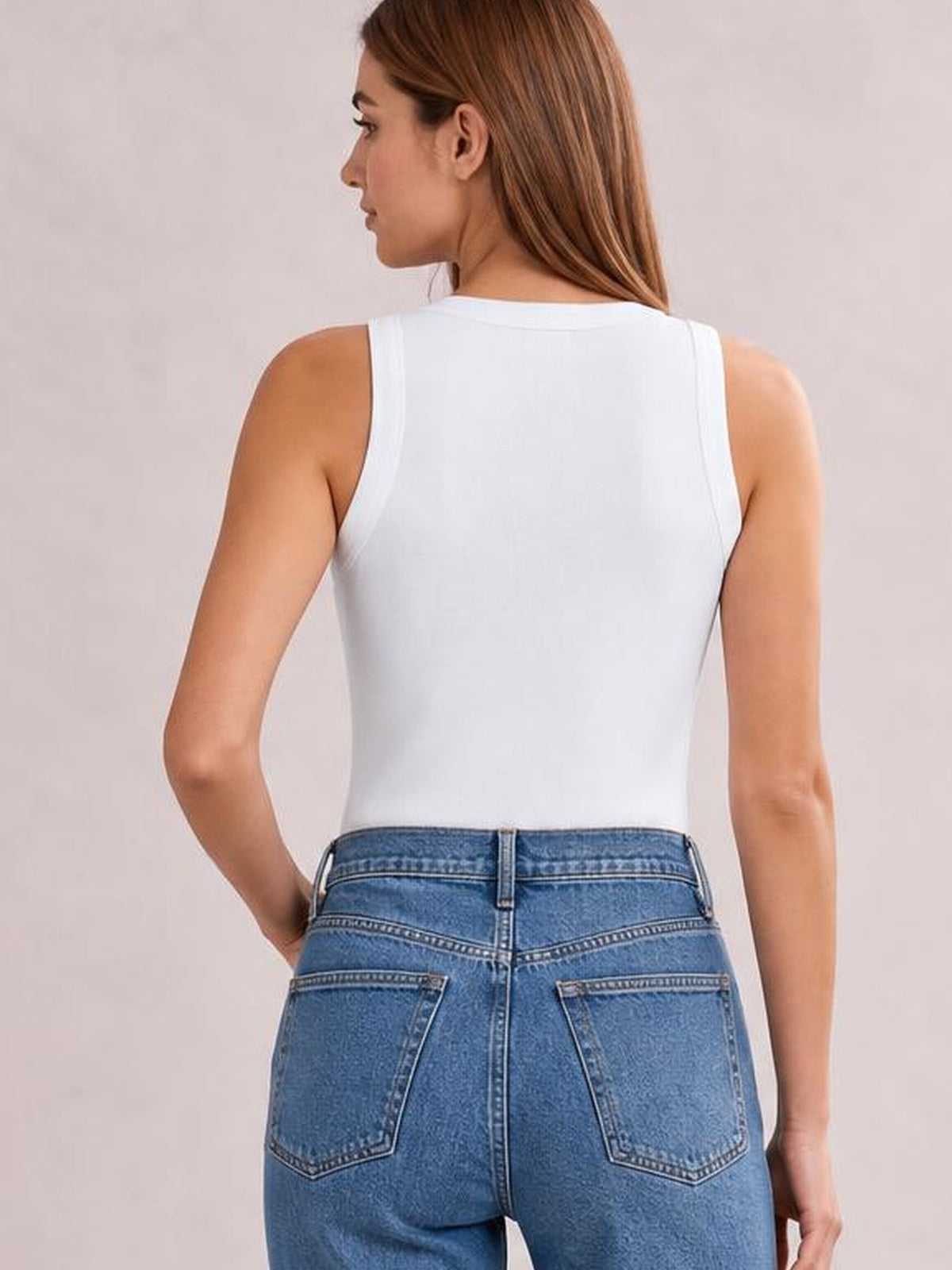 Model shown from behind in a white solid round neck tank with sleeveless cut, clean construction, and a fitted silhouette as one hand rests at her hip.